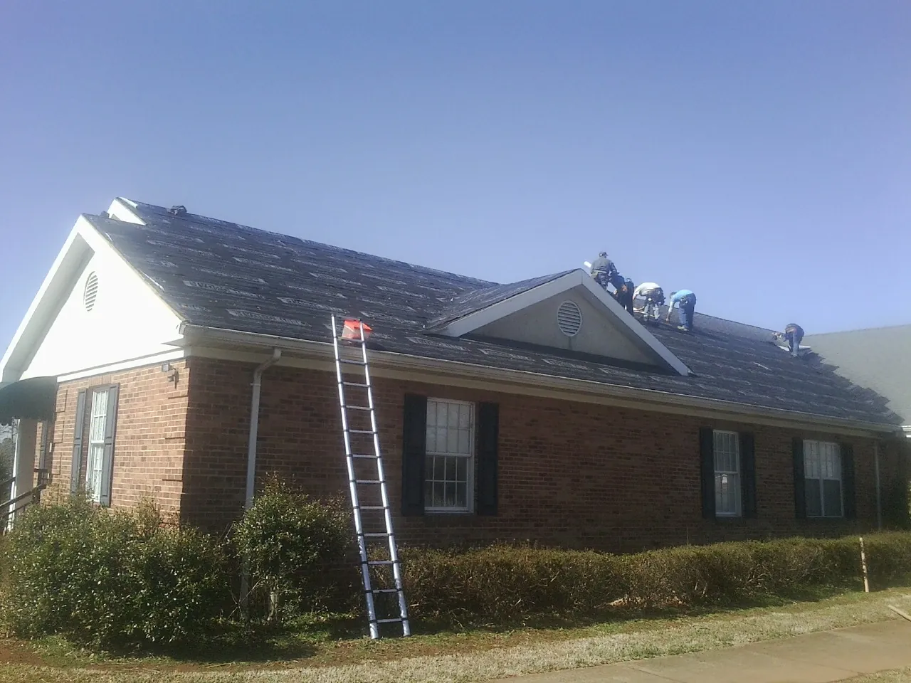Crew tearing shingles off a brick ranch roof with a ladder set — Metal Roof Repair work by our crew in Mc Alisterville
