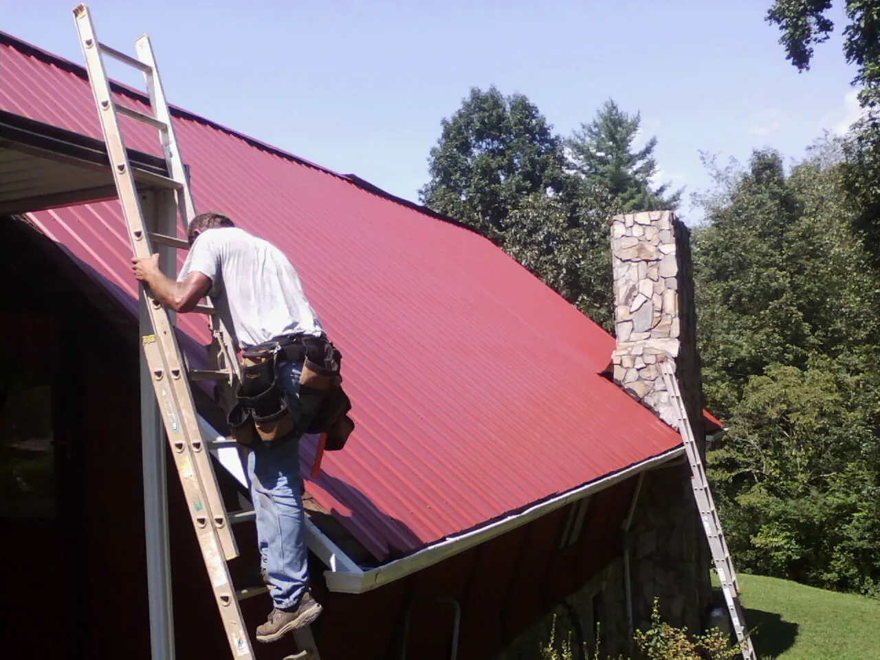 Roofer on a ladder beside a red metal roof and stone chimney — Soffit & Fascia Repair by Roofing Brothers in Mc Alisterville